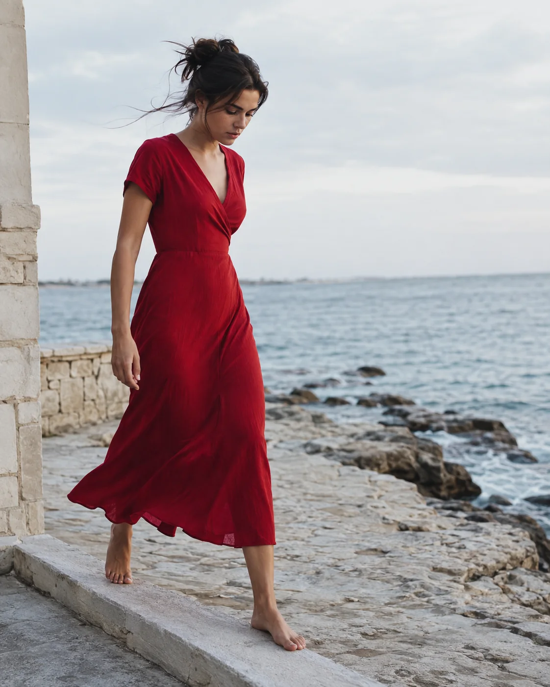 A dark-haired female character in a red dress, barefoot on a stone threshold by the sea, looking down as the wind moves the fabric.